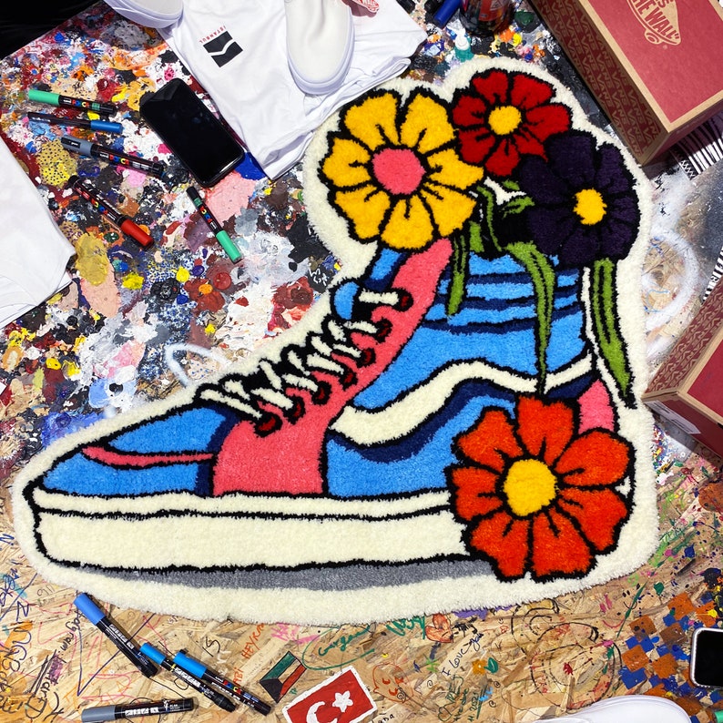 Może przedstawiać: Wysokie niebiesko-r&oacute;żowe buty sportowe z białymi sznurowadłami i białą podeszwą są przedstawione w projekcie dywanu. Buty są otoczone kolorowymi kwiatami w odcieniach czerwieni, ż&oacute;łci i fioletu.