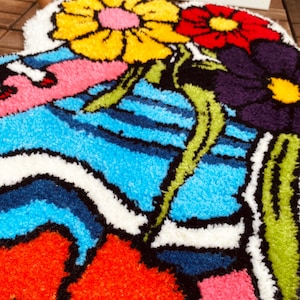 Może przedstawiać: Kolorowy dywan z kwiatowym wzorem. Dywan przedstawia duży ż&oacute;łty kwiat z czerwonymi i białymi akcentami, mniejszy czerwony kwiat z białymi akcentami i fioletowy kwiat z białymi akcentami. Dywan ma r&oacute;wnież wz&oacute;r w niebieskie i białe pasy.