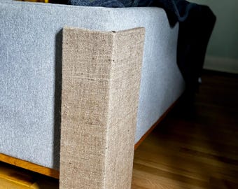 May include: A corner of a light gray upholstered sofa with a natural burlap fabric accent. The sofa has a wooden base and is set on a hardwood floor. A dark blue blanket is draped over the back of the sofa.