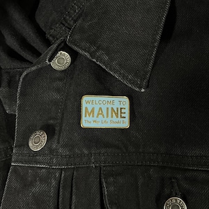 May include: Gold-colored enamel pin with a blue background. The pin says "Welcome to Maine, The Way Life Should Be".