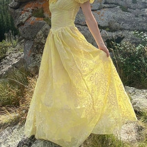 May include: A yellow, sheer, long, floral-patterned dress with a ruffled neckline and short sleeves. The dress is flowing and has a vintage style.