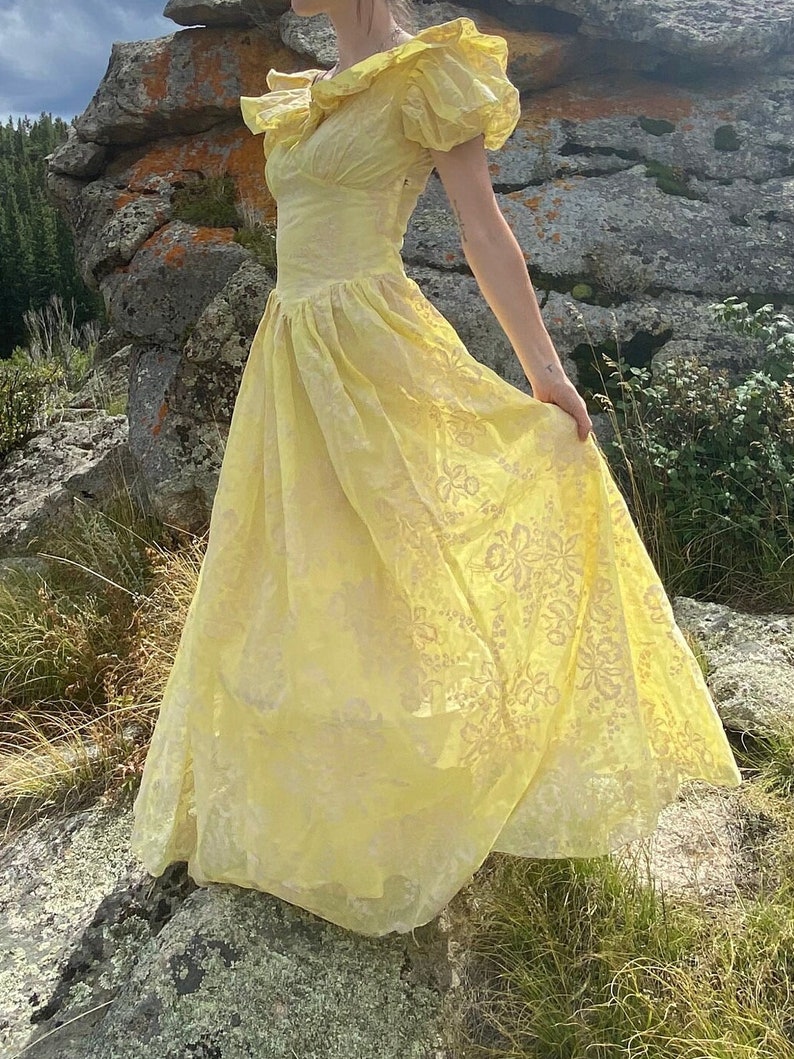 May include: A yellow, sheer, long, floral-patterned dress with a ruffled neckline and short sleeves. The dress is flowing and has a vintage style.