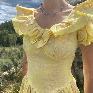 May include: A yellow dress with a floral pattern and a bow at the neckline. The dress has a fitted bodice and a full skirt. The model is wearing a silver necklace with bow charms.