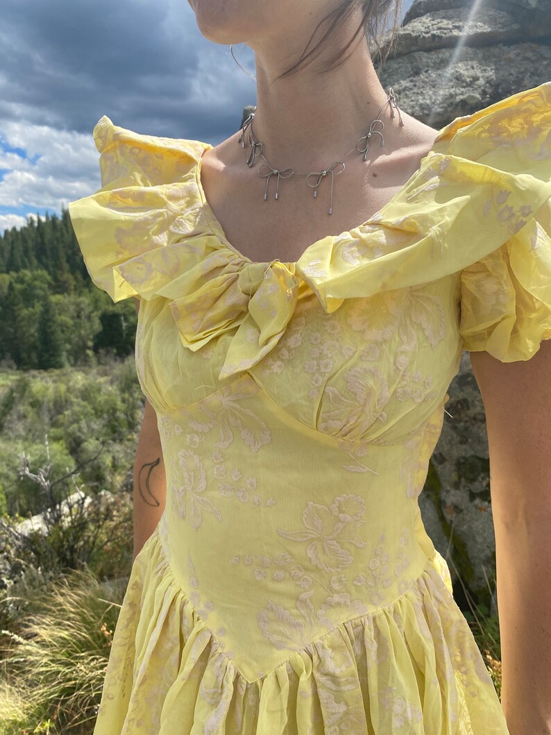 May include: A yellow dress with a floral pattern and a bow at the neckline. The dress has a fitted bodice and a full skirt. The model is wearing a silver necklace with bow charms.