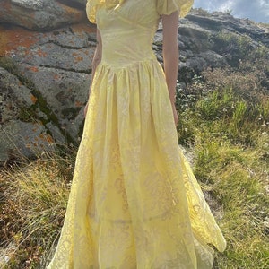 May include: A yellow vintage prom dress with a ruffled neckline and short sleeves. The dress has a floral pattern and a full skirt.