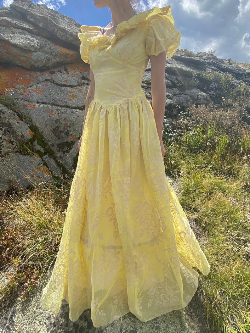 May include: A yellow vintage prom dress with a ruffled neckline and short sleeves. The dress has a floral pattern and a full skirt.