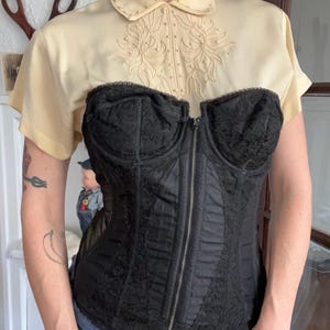 May include: A black lace corset with a front zipper is worn over a light yellow collared shirt with floral embroidery. The corset has a structured design and is paired with blue jeans.