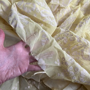 May include: A yellow sheer fabric with a white floral pattern. The fabric is slightly wrinkled and has a frayed edge.