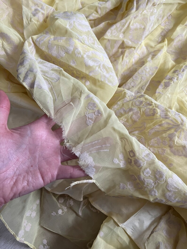 May include: A yellow sheer fabric with a white floral pattern. The fabric is slightly wrinkled and has a frayed edge.