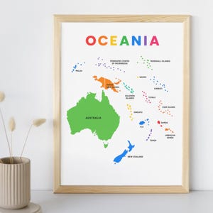 Op de afbeelding: Een ingelijste Oceania-kaartprint met een licht houten frame. De kaart toont het woord "OCEANIA" in regenboogkleuren. Australië is groen en Nieuw-Zeeland blauw. Andere eilanden zijn gekleurd in oranje, paars en geel, met landnamen.