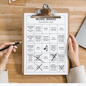 Puede incluir: Un portapapeles con una hoja de juego "MUSIC BINGO" impresa con &eacute;xitos de fiesta de la d&eacute;cada de 2010. La hoja est&aacute; parcialmente marcada con cruces de l&aacute;piz. Una mano sostiene un l&aacute;piz, lista para marcar la hoja. Se ve un port&aacute;til.