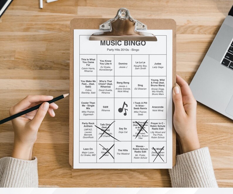 Puede incluir: Un portapapeles con una hoja de juego "MUSIC BINGO" impresa con &eacute;xitos de fiesta de la d&eacute;cada de 2010. La hoja est&aacute; parcialmente marcada con cruces de l&aacute;piz. Una mano sostiene un l&aacute;piz, lista para marcar la hoja. Se ve un port&aacute;til.