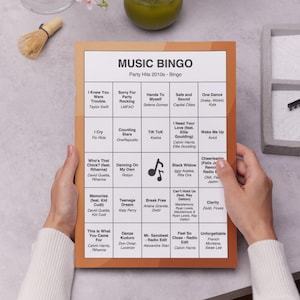 Puede incluir: Una tarjeta de juego Music Bingo con un borde marr&oacute;n, con t&iacute;tulos de canciones y artistas de la d&eacute;cada de 2010. La tarjeta est&aacute; en manos de una persona. El t&iacute;tulo "MUSIC BINGO" est&aacute; en la parte superior.