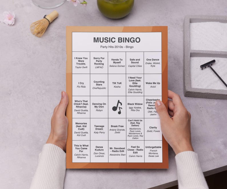 Puede incluir: Una tarjeta de juego Music Bingo con un borde marr&oacute;n, con t&iacute;tulos de canciones y artistas de la d&eacute;cada de 2010. La tarjeta est&aacute; en manos de una persona. El t&iacute;tulo "MUSIC BINGO" est&aacute; en la parte superior.
