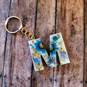 May include: A white resin letter M keychain with a gold chain. The letter is filled with blue and white pressed flowers and gold leaf.