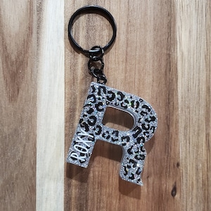 May include: A silver and black glitter keychain with the letter R in a leopard print design. The keychain has a black metal ring and a silver chain. The letter R has the word "ROXY" written on it in white.