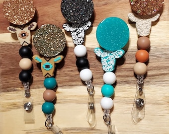 May include: Five badge reels with glittery circular tops and decorative cow skull accents. The reels feature various bead colors, including black, teal, and tan, with clear plastic clips for attaching to clothing.
