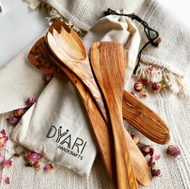 Puede incluir: Conjunto de cuatro utensilios de cocina de madera de olivo, que incluyen una esp&aacute;tula, una cuchara para servir y dos cucharas m&aacute;s peque&ntilde;as. Los utensilios est&aacute;n dispuestos sobre un fondo de tela blanca con el nombre de la marca "Diyari Handcrafts" impreso en una peque&ntilde;a bolsa blanca.
