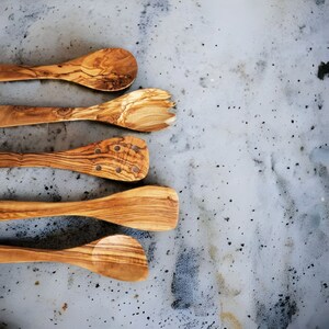 Puede incluir: Cinco utensilios de cocina de madera, incluyendo una cuchara ranurada, una cuchara y una esp&aacute;tula, est&aacute;n dispuestos verticalmente sobre una superficie texturizada gris claro.