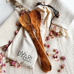 May include: A set of two olive wood salad servers in a natural canvas bag with the text "DYARI HANDCRAFTS". The servers are on a white background with dried pink rose buds.