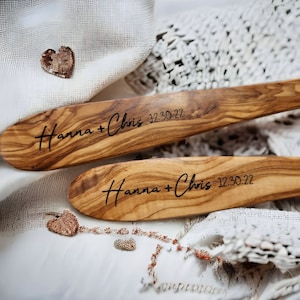 Puede incluir: Dos cucharas de ensalada de madera con los nombres "Hanna + Chris" y la fecha "12.30.22" grabados. Las cucharas est&aacute;n sobre una tela blanca de crochet.