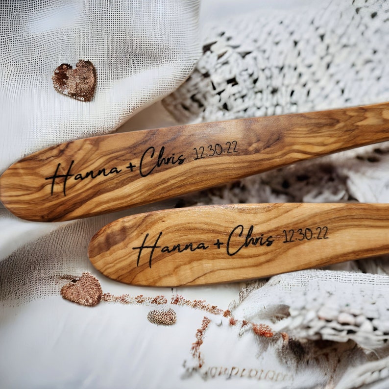 Puede incluir: Dos cucharas de ensalada de madera con los nombres "Hanna + Chris" y la fecha "12.30.22" grabados. Las cucharas est&aacute;n sobre una tela blanca de crochet.