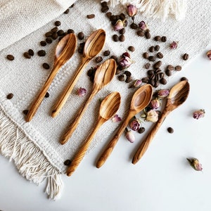 May include: A set of six wooden spoons with natural wood grain. The spoons are arranged on a white fabric background with coffee beans and dried flowers.