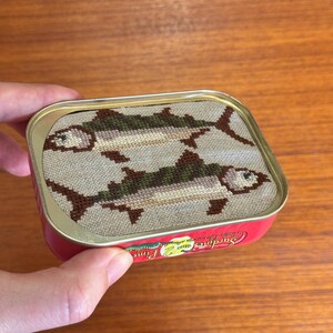 Może przedstawiać: Czerwono-biała puszka z haftem krzyżykowym przedstawiającym dwie ryby na pokrywce. Tekst na puszce brzmi "Sardines Fine" i "Jakość Gwarantowana".