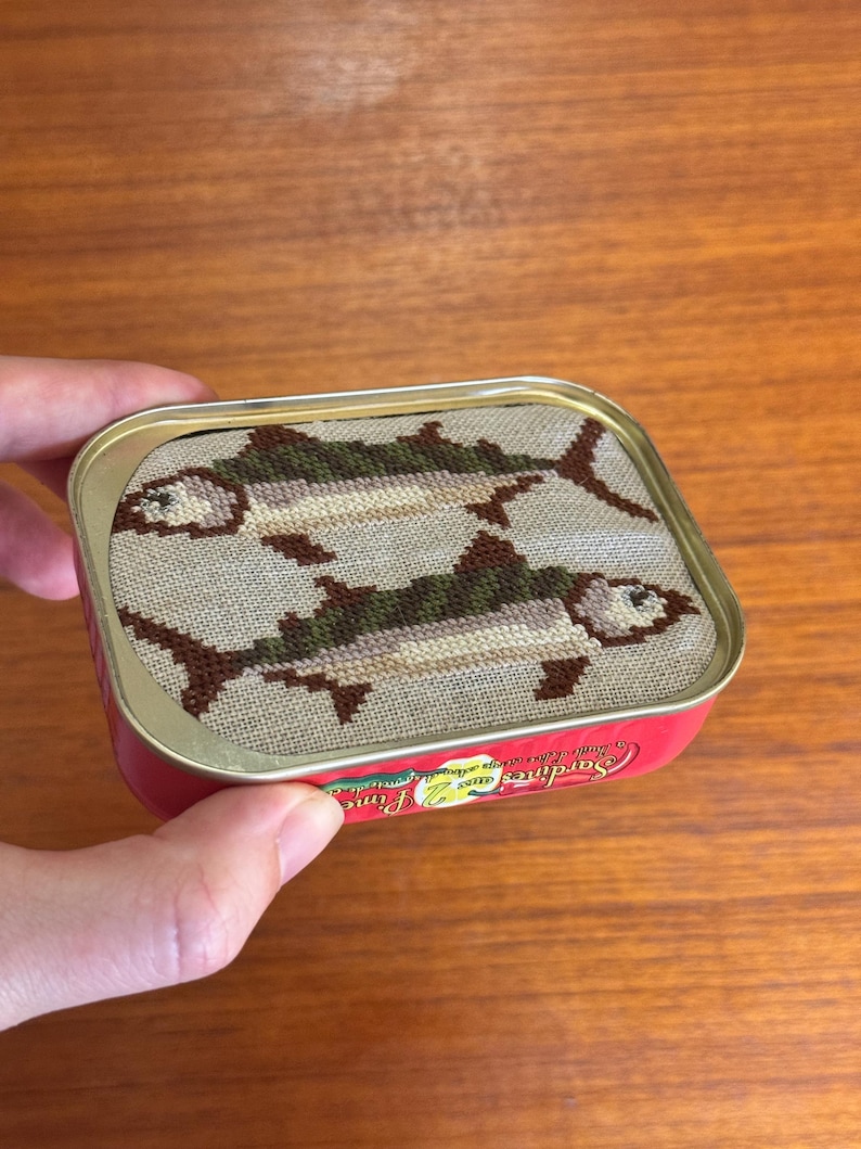 Może przedstawiać: Czerwono-biała puszka z haftem krzyżykowym przedstawiającym dwie ryby na pokrywce. Tekst na puszce brzmi "Sardines Fine" i "Jakość Gwarantowana".