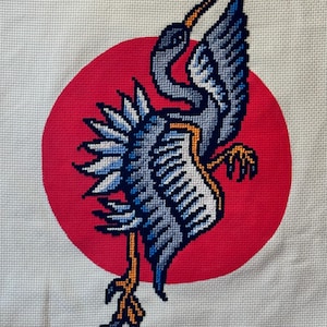May include: A cross-stitch embroidery of a blue and grey crane with a long orange beak, standing on a red circle.