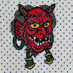 Puede incluir: Bordado de punto de cruz de un demonio oni rojo y negro con detalles verdes y amarillos. El oni lleva una máscara de demonio japonesa tradicional con cuernos y colmillos. El fondo es una tela blanca con lunares azules.