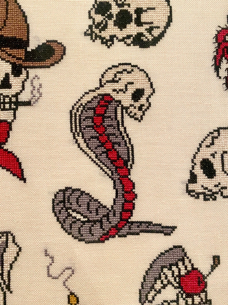 May include: Cross-stitch embroidery featuring a gray and red cobra with a skull head. The cobra is curled up with its tail pointing to the right. The background is a white fabric.