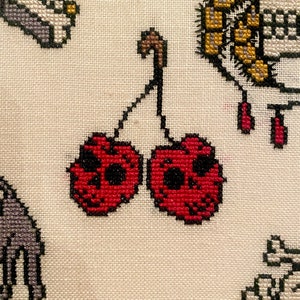 Peut inclure: Broderie au point de croix de deux cerises rouges avec des visages de crânes noirs. Les cerises sont reliées par une tige brune.