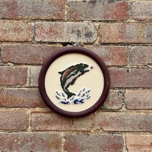 May include: A round, framed cross-stitch embroidery featuring a leaping fish. The fish is primarily black with red and green accents, jumping out of blue water. The embroidery is set against a cream-colored background and framed in a dark wood hoop.