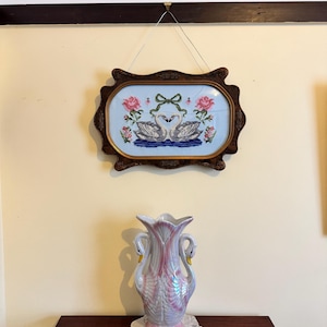 May include: A framed cross-stitch featuring two swans facing each other, with pink roses and a green bow. Below, a decorative vase with swan-shaped handles and a pink and white iridescent finish sits on a dark wooden table.