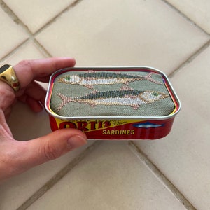 Może przedstawiać: Mała, prostokątna puszka z czerwoną i ż&oacute;łtą etykietą z napisem "Ortiz Sardines". Puszka jest ozdobiona zielonym, materiałowym wierzchem z haftem krzyżykowym przedstawiającym dwie ryby.