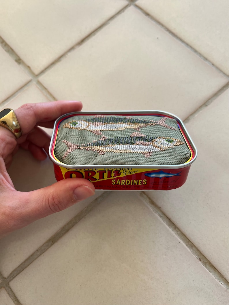 Może przedstawiać: Mała, prostokątna puszka z czerwoną i ż&oacute;łtą etykietą z napisem "Ortiz Sardines". Puszka jest ozdobiona zielonym, materiałowym wierzchem z haftem krzyżykowym przedstawiającym dwie ryby.