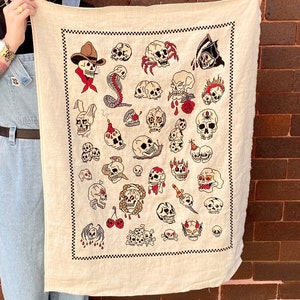 May include: A white fabric wall tapestry with a black checkered border. The tapestry features a variety of colorful embroidered skull designs, including a cowboy skull, a snake skull, and a skull with a crown.