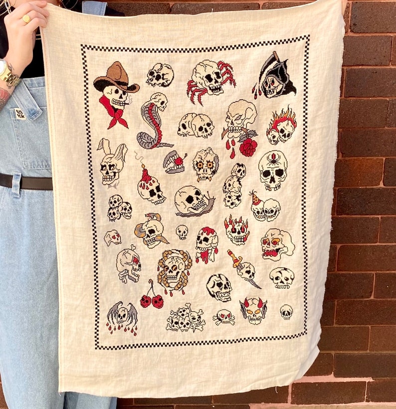 May include: A white fabric wall tapestry with a black checkered border. The tapestry features a variety of colorful embroidered skull designs, including a cowboy skull, a snake skull, and a skull with a crown.