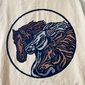 May include: A cross-stitch embroidery of three horse heads in a circle. The horses are brown, gray, and black with brown, gray, and white markings. The background is a cream color.