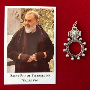 Könnte beinhalten: Eine rechteckige Gebetskarte mit einem Porträt von Pater Pio von Pietrelcina und dem Text "Saint Pio of Pietrelcina" sowie "Padre Pio" darunter, neben einem silberfarbenen Metallanhänger in Form eines kreisförmigen Rosenkranzes mit Porträt.