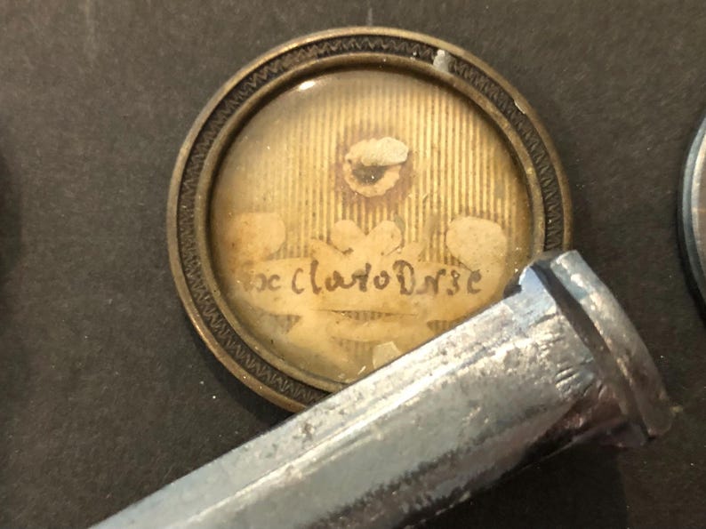 May include: A small, round, gold-coloured locket with a glass cover. The locket contains a faded image of a flower and the text "Ex. Claudio Dn.e".