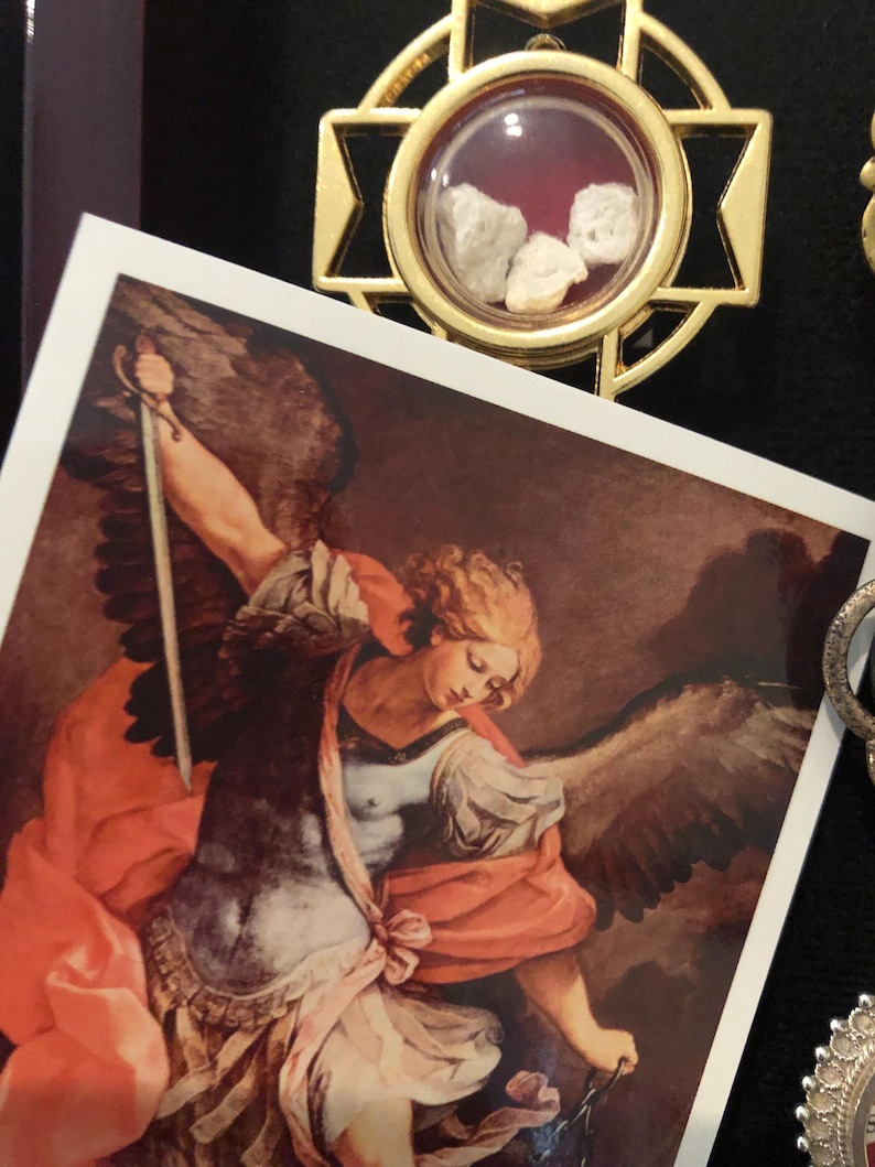 以下が含まれることがあります： 金色の十字型のフレームが付いた宗教的なペンダント。透明なコンパートメントには、3つの小さな淡い色の石が入っています。剣と翼を持つ天使のプリントが前景にあります。