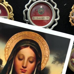 Op de afbeelding: Een close-up van een zilveren ingelijste relikwie met een rode achtergrond en de tekst "S. Lucidi M." erin. De relikwie ligt op een wit oppervlak met een schilderij van de Maagd Maria op de achtergrond.