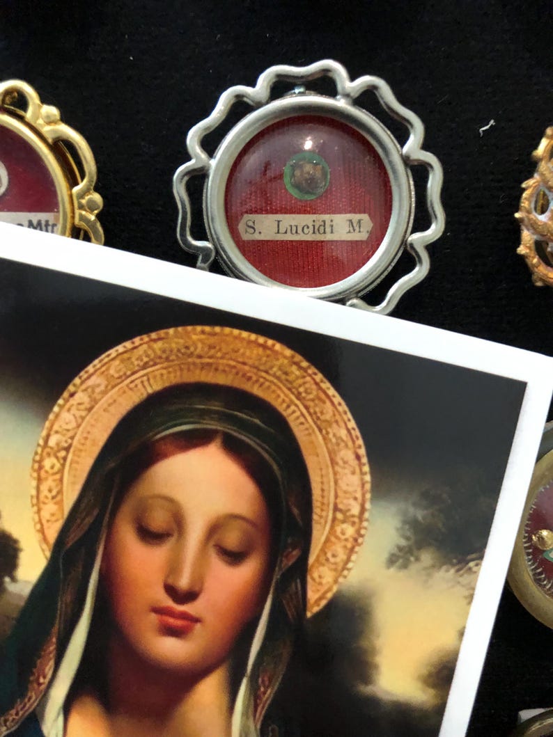 Op de afbeelding: Een close-up van een zilveren ingelijste relikwie met een rode achtergrond en de tekst "S. Lucidi M." erin. De relikwie ligt op een wit oppervlak met een schilderij van de Maagd Maria op de achtergrond.