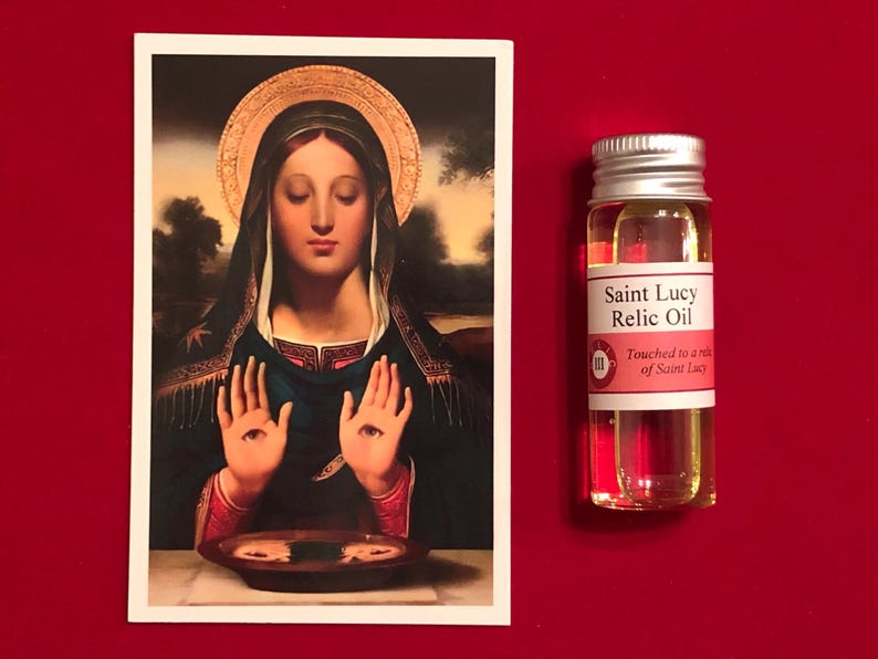 Op de afbeelding: Een klein glazen flesje met reliek-olie van Sint Lucia. De fles heeft een wit etiket met de tekst "Saint Lucy Relic Oil" en "Touched to a relic of Saint Lucy".