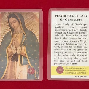 May include: A prayer card featuring an image of Our Lady of Guadalupe, with a teal cloak and clasped hands. The reverse side contains a prayer and information about a relic. The card is encased in a clear plastic sleeve.