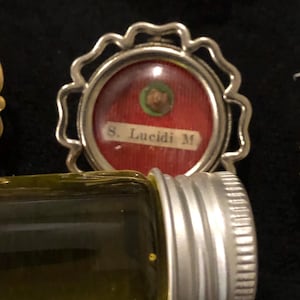 Op de afbeelding: Een kleine, ronde reliek met een zilveren frame, een rode achtergrond en een kleine groene cirkel met een vervaagde afbeelding. De tekst "S. Lucidi M." is in wit op de rode achtergrond gedrukt.