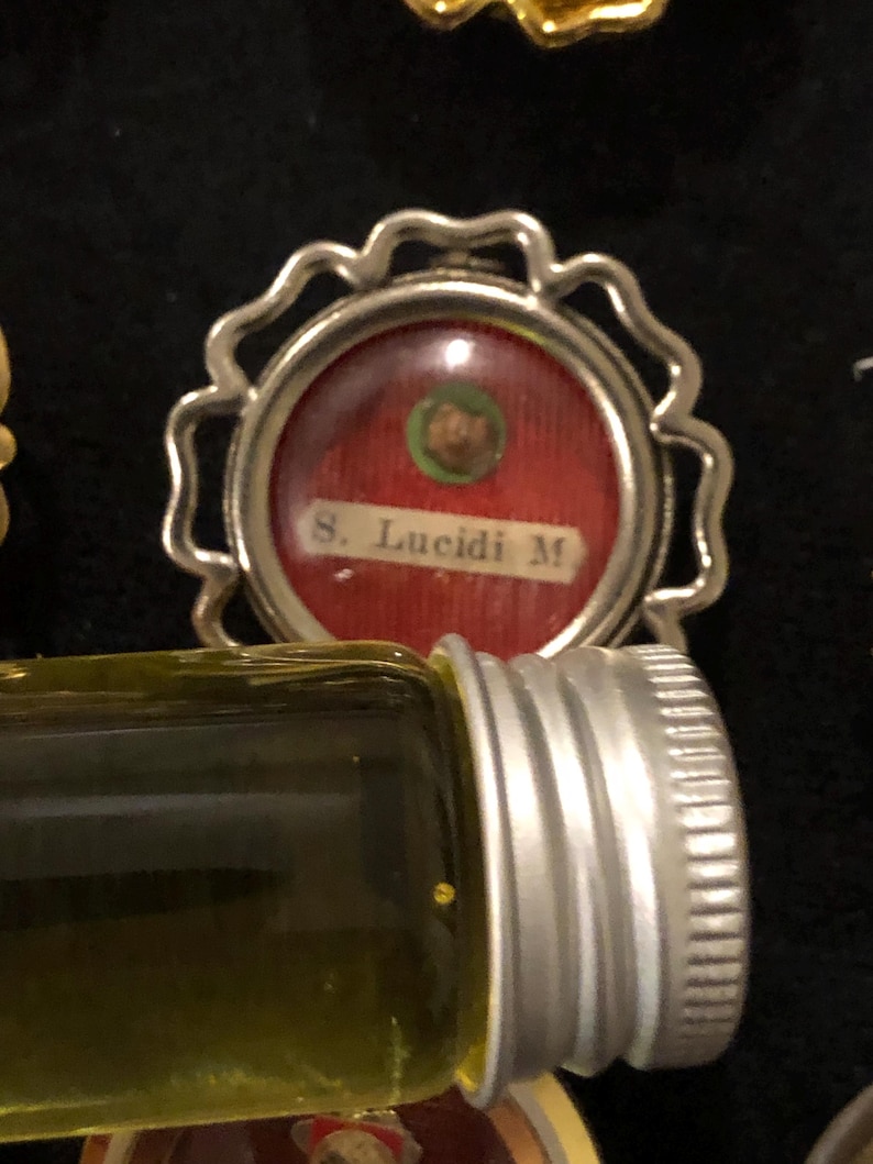 Op de afbeelding: Een kleine, ronde reliek met een zilveren frame, een rode achtergrond en een kleine groene cirkel met een vervaagde afbeelding. De tekst "S. Lucidi M." is in wit op de rode achtergrond gedrukt.