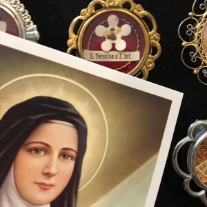 Puede incluir: Una reliquia religiosa vintage con una imagen enmarcada en oro de una flor blanca sobre fondo rojo. La imagen est&aacute; rodeada por un marco de filigrana dorada e incluye el texto "S. Teresiae a J. Inf."
